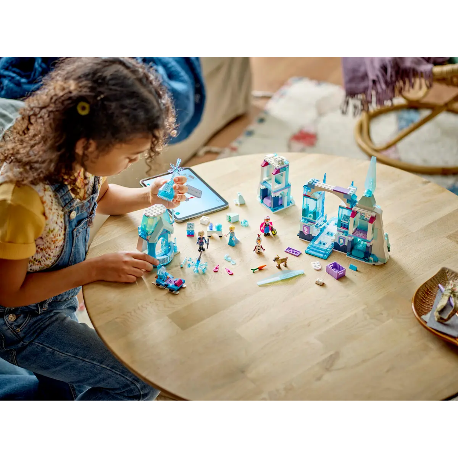Een meisje speelt met een Lego-set van een kasteel op een houten tafel. De set bevat figuren en accessoires. Een tablet is in de buurt.