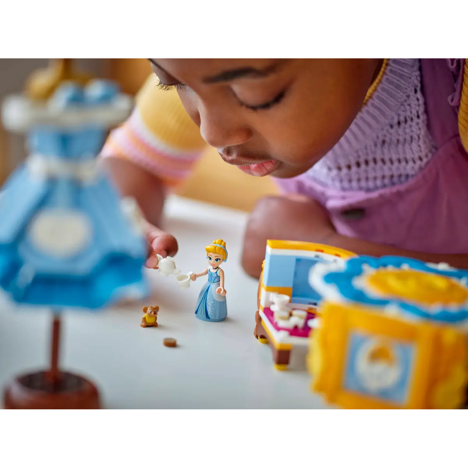 A child playing with Cinderella Lego figures. A blue dress, a bed, a small bear, and a teapot are on the table.