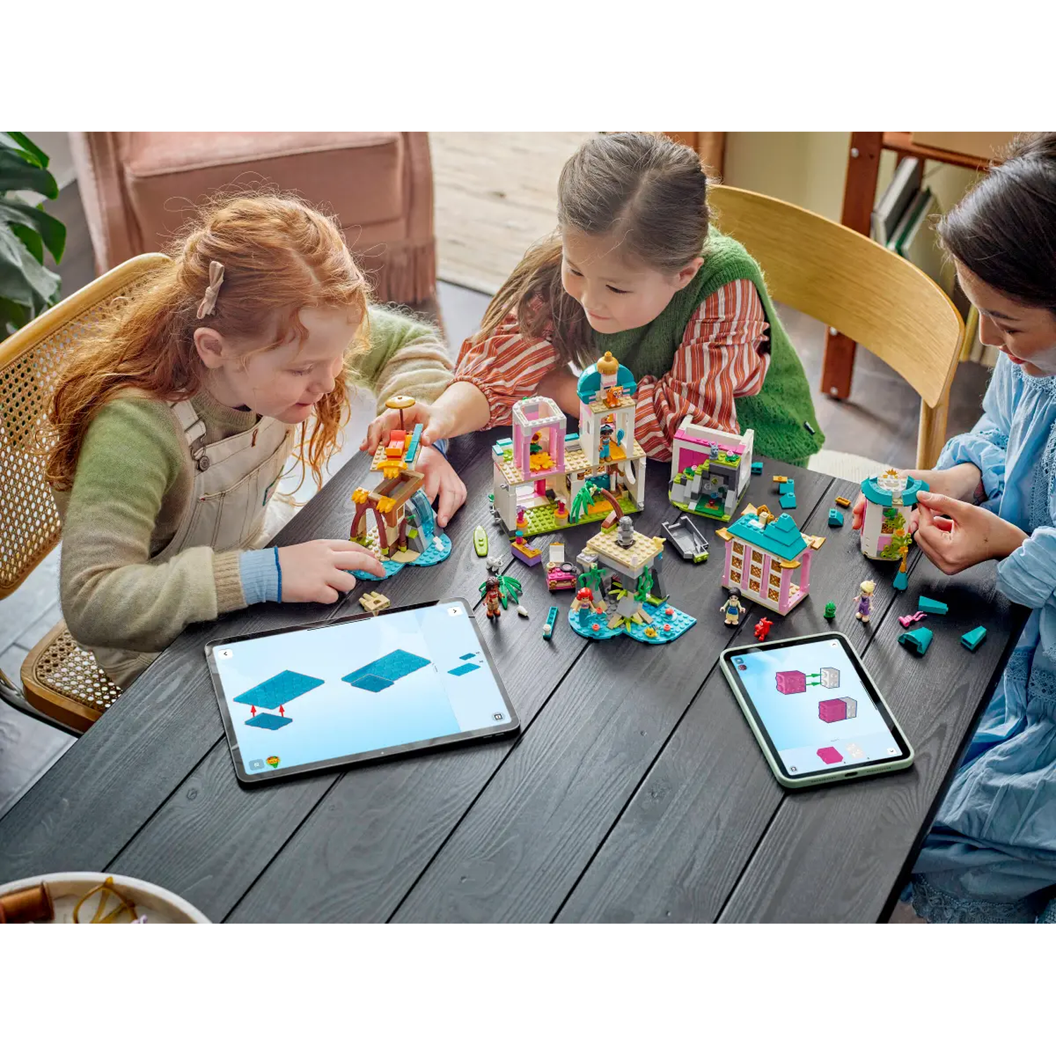 Drie meisjes bouwen Lego-sets op een zwarte tafel. Tablets tonen instructies. Roze, groene en witte Lego-structuren zijn zichtbaar.