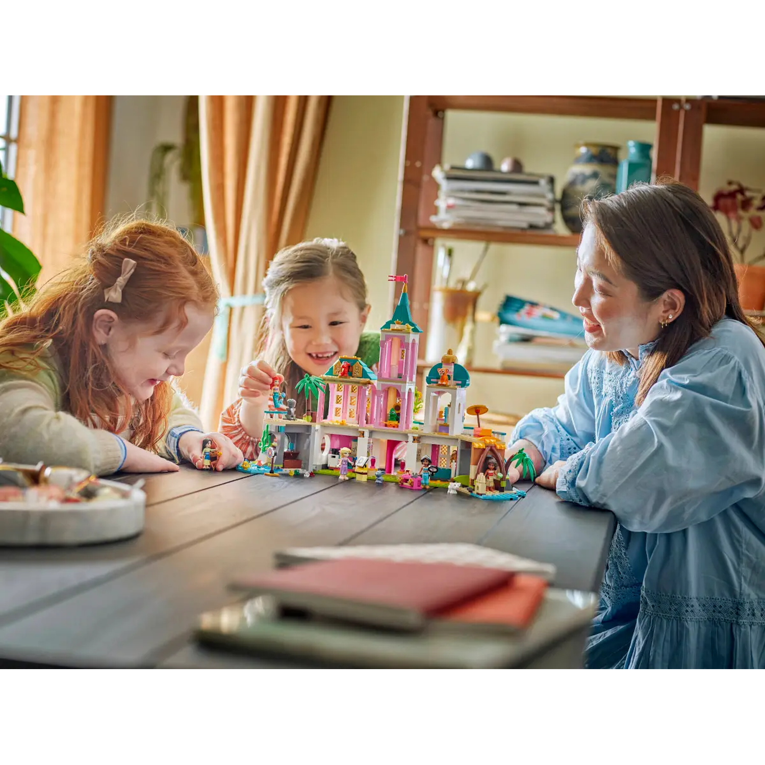 Drie mensen rond een tafel die met een Lego-set spelen. Een roze en wit kasteel staat in het midden. Ze lachen.
