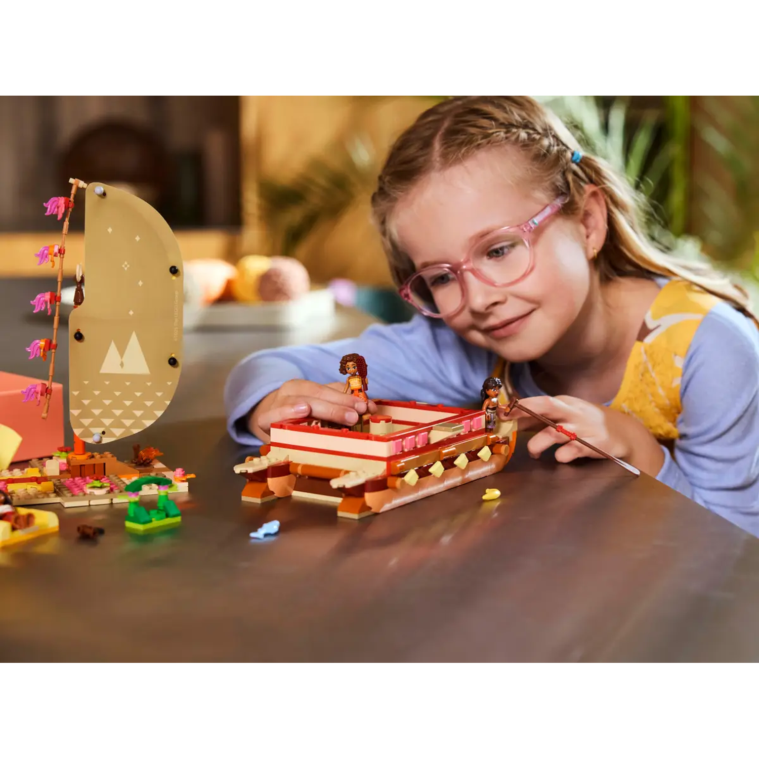 A girl with glasses looks at a Lego boat with figures. A sail and other Lego pieces are on the table.