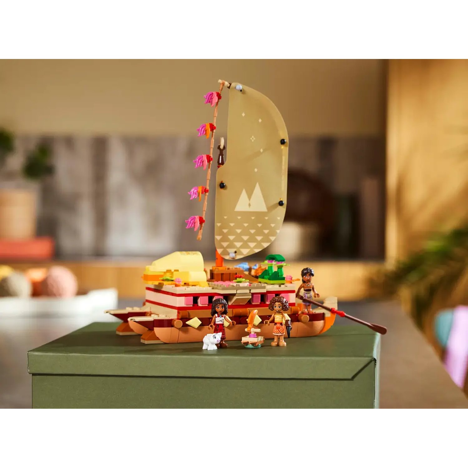 A Lego boat with a tan sail, featuring Lego figures. The boat is on a green box, with a blurred background of a kitchen.
