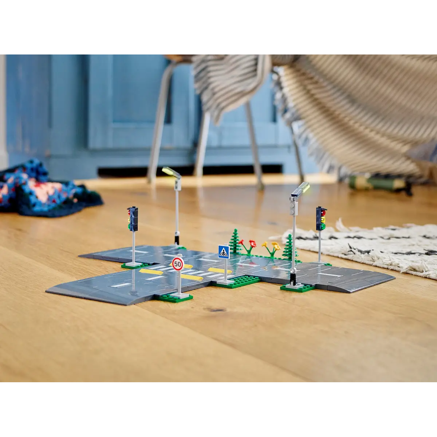 A Lego city scene on wooden floor. It includes roads, traffic lights, signs, and decorative plants. Background has furniture.