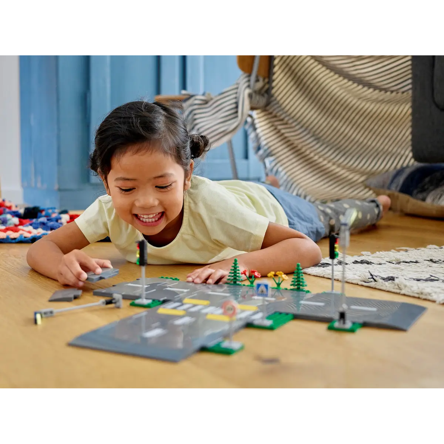 A girl plays with a Lego city set on the floor. She is smiling. Road pieces, trees, and traffic lights are visible.