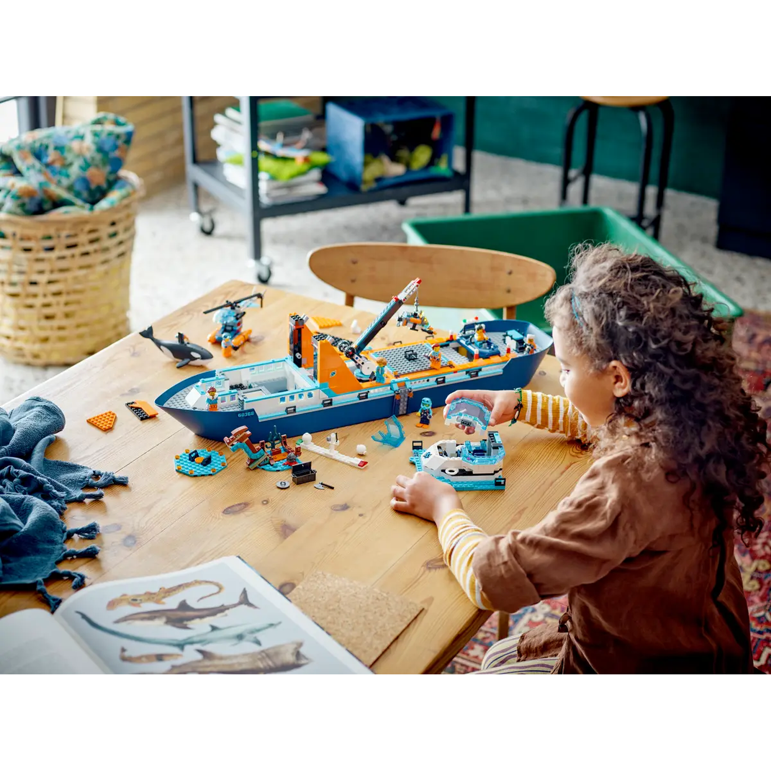 A child is playing with a Lego set on a wooden table. A boat, helicopter and shark models are present. A book with shark images is open.