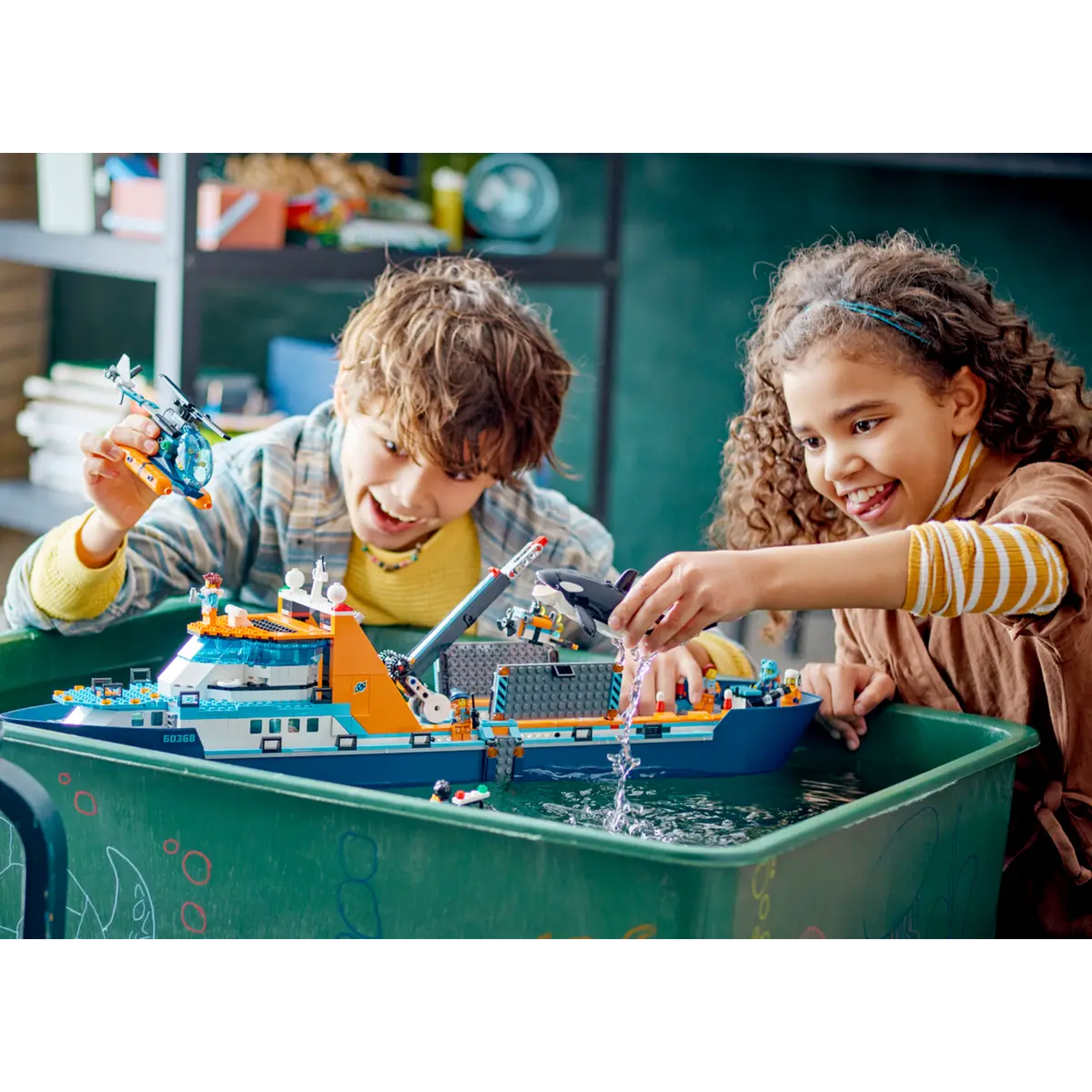 Two children playing with a Lego ship in a green bin filled with water. One holds a helicopter, the other pours water.