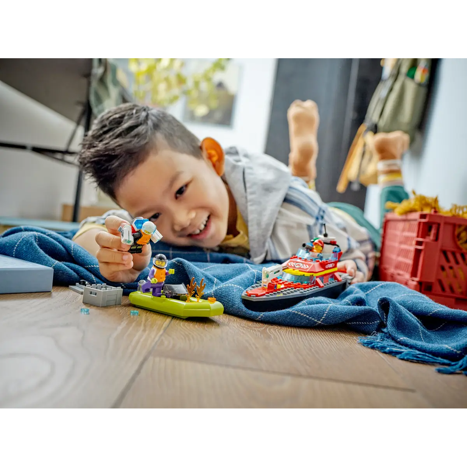 A smiling boy plays with Lego boats and figures on a blue blanket. A red crate is in the background.