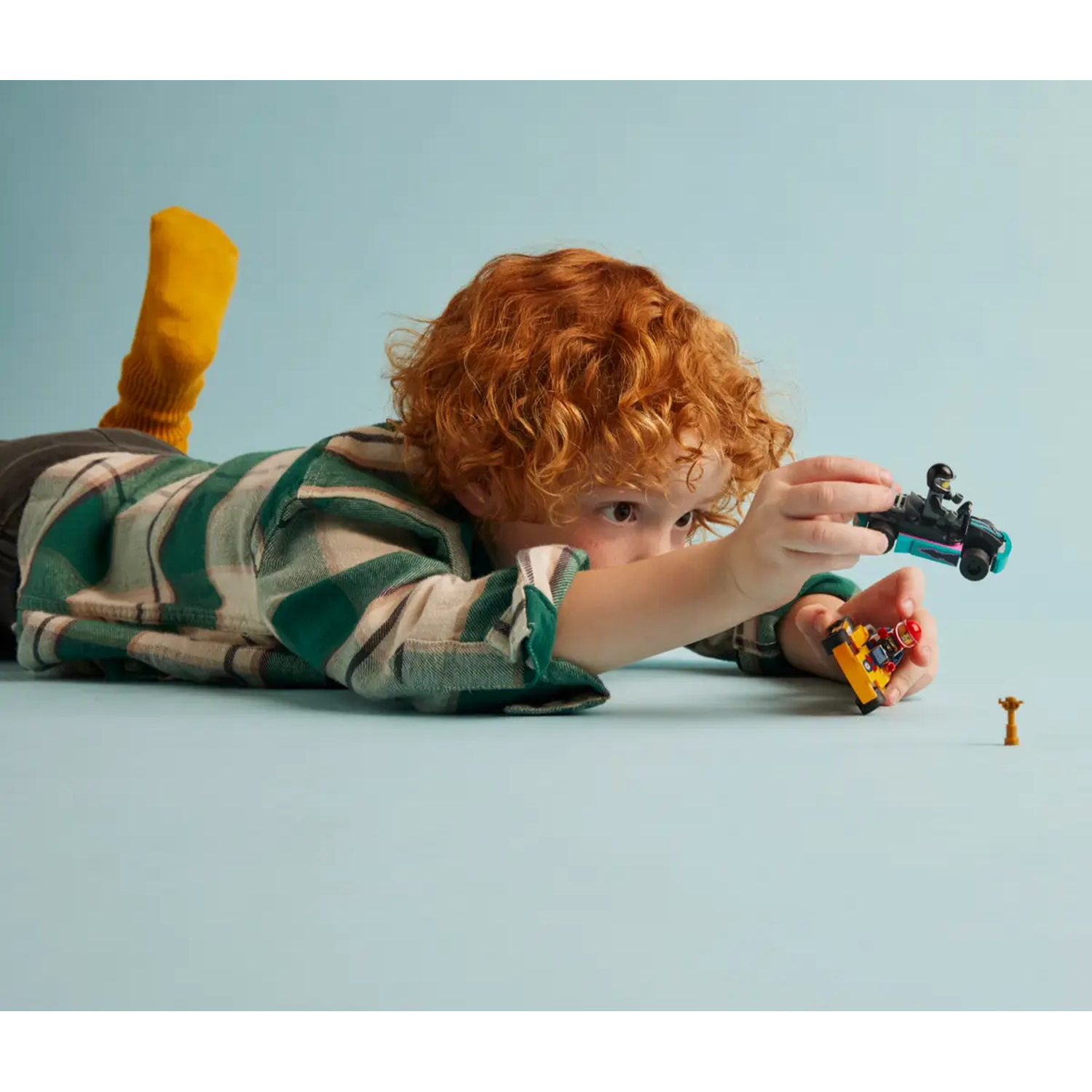A child plays with toy cars on a blue surface. The child is wearing a green shirt and a yellow sock. The toy cars are black and yellow.