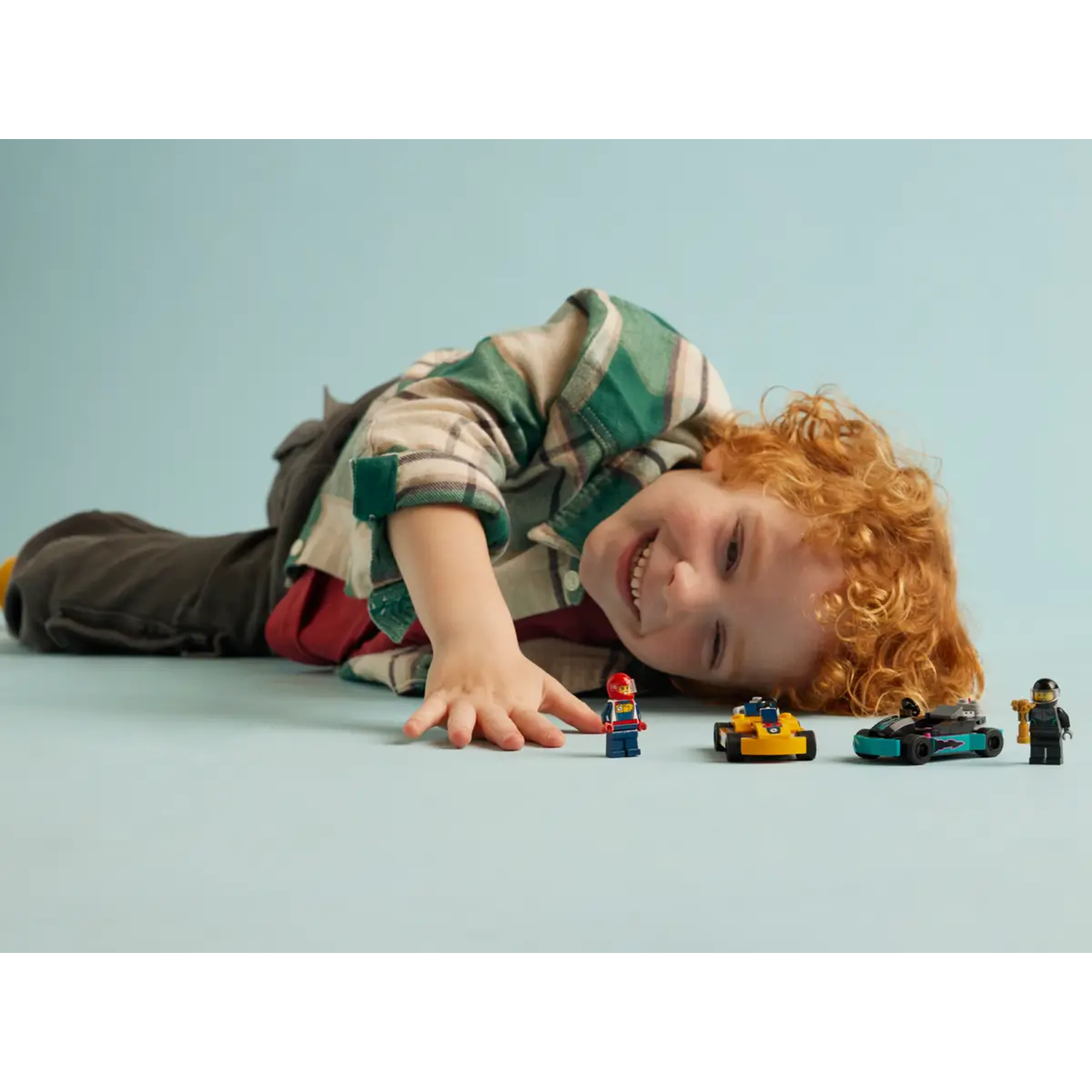 A child smiles, reaching towards toy race cars and figures. Background is light blue.