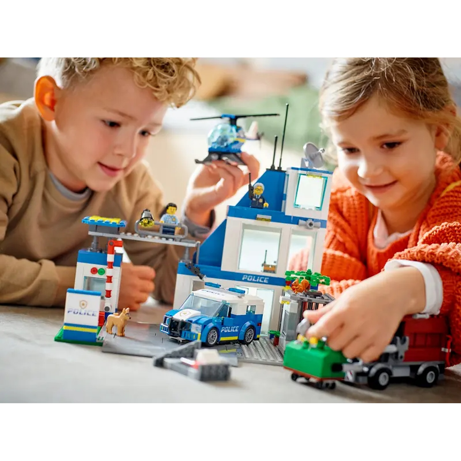 Two children playing with a Lego police station, including a police car, helicopter, and figures. The scene is bright and colorful.