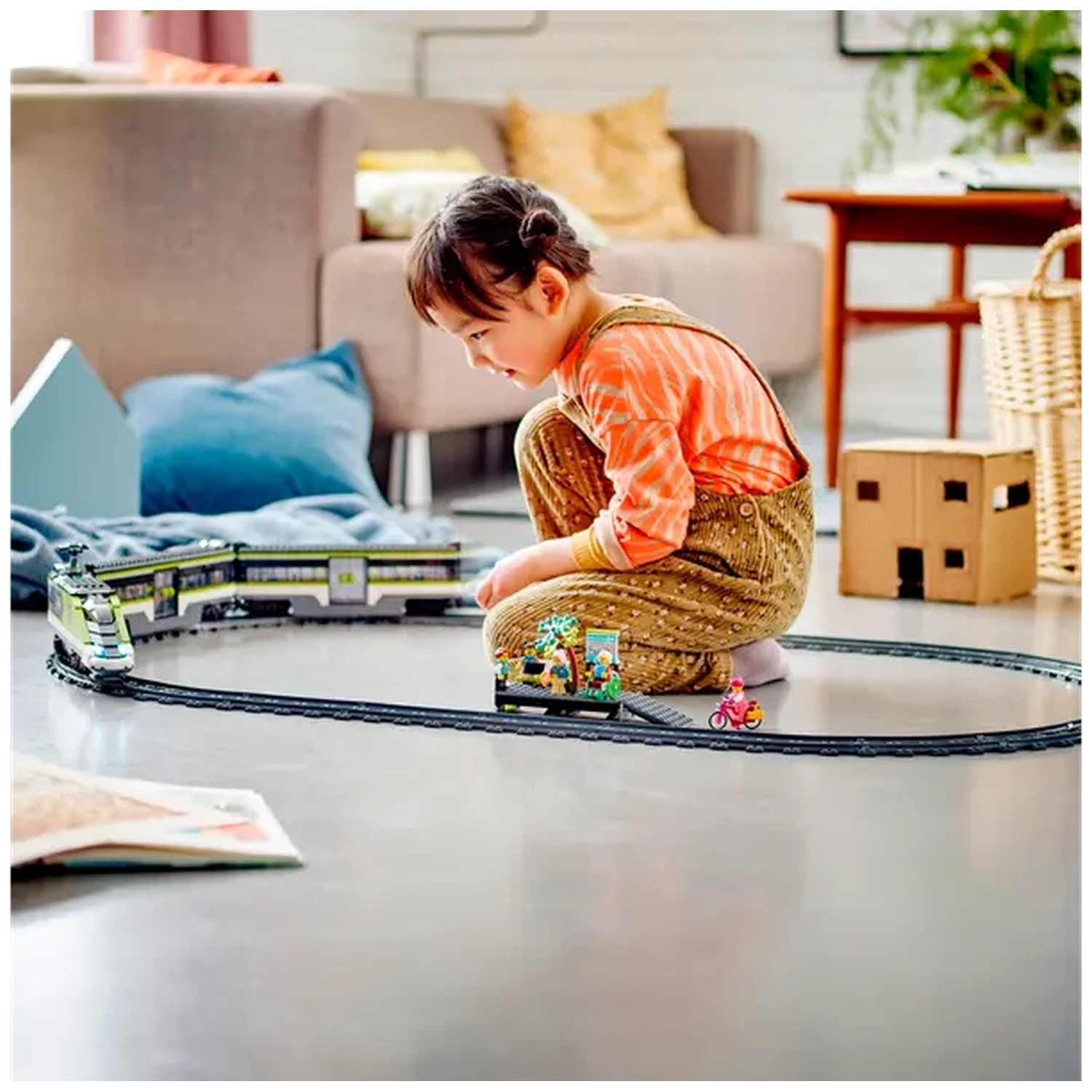 A child plays with a toy train set on the floor. A toy train, a small house and a bicycle are also visible.