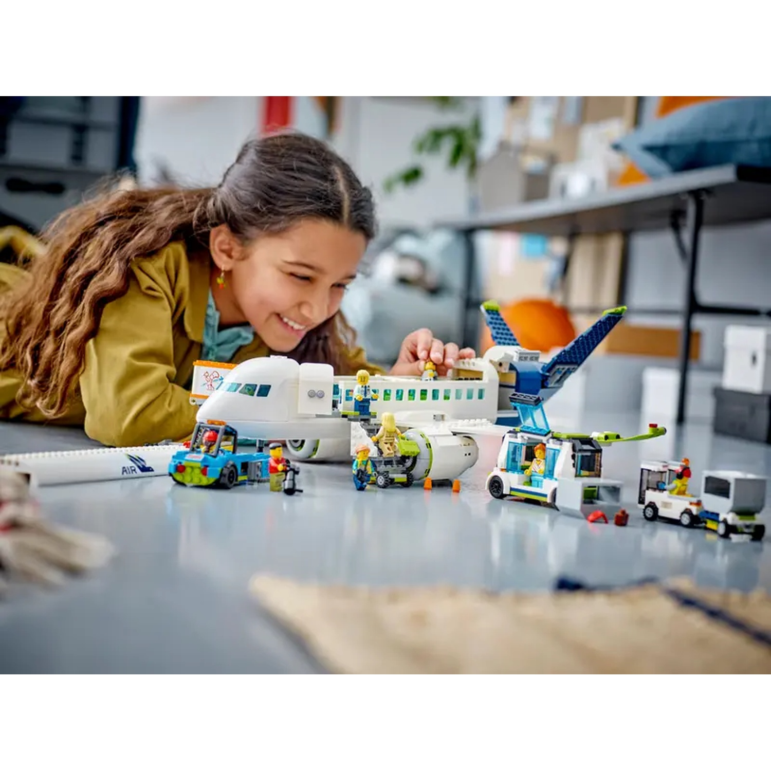 A girl smiles at a LEGO airport scene. A white plane, blue and green vehicles, and figures are on a gray floor.