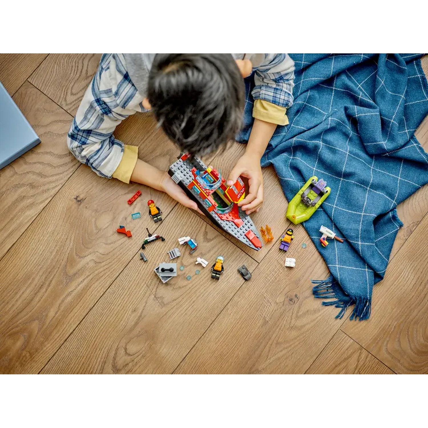 Un enfant joue avec des jouets de bateaux Lego sur un sol en bois, avec une couverture bleue et un livre. Des figurines Lego sont éparpillées.