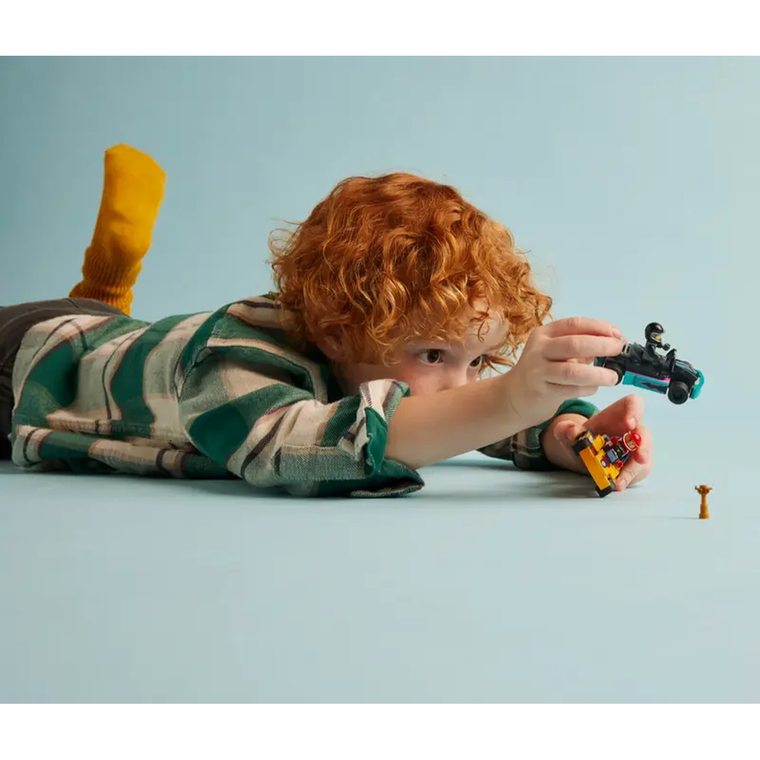 A child with red hair plays with toy cars. He is lying on the floor, wearing a striped shirt and a yellow sock. The background is blue.