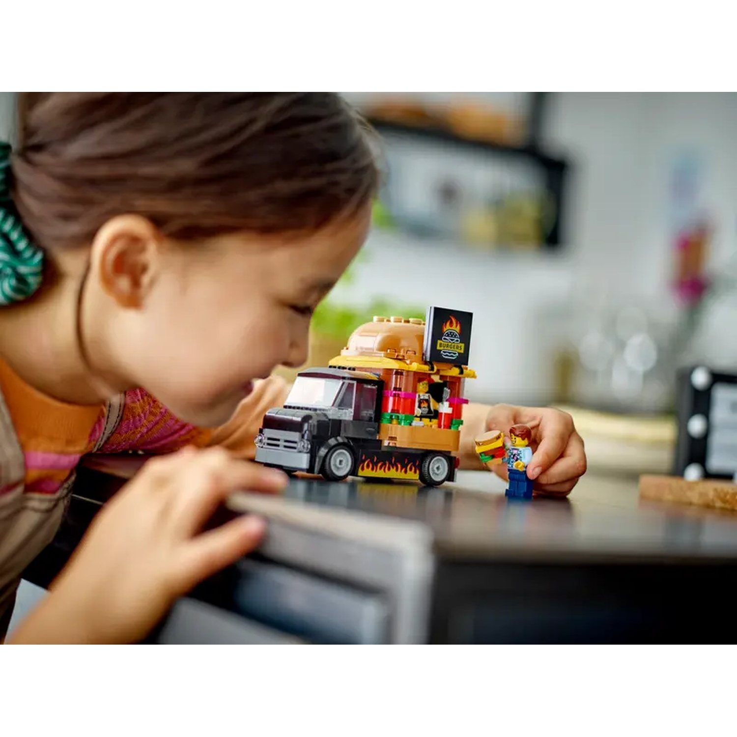 A girl looks at a Lego burger truck. A Lego figure holds a burger. The truck has a sign that says 'Burgers'.