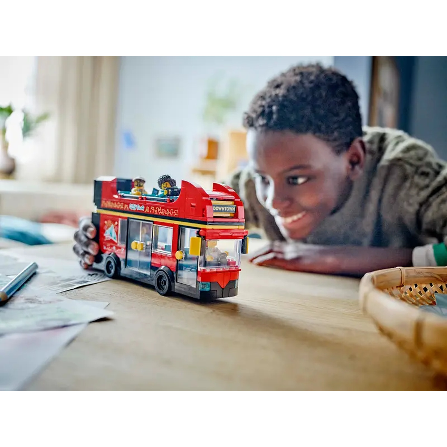 A smiling boy looks at a red Lego double-decker bus on a wooden table. The bus has small figures on top.
