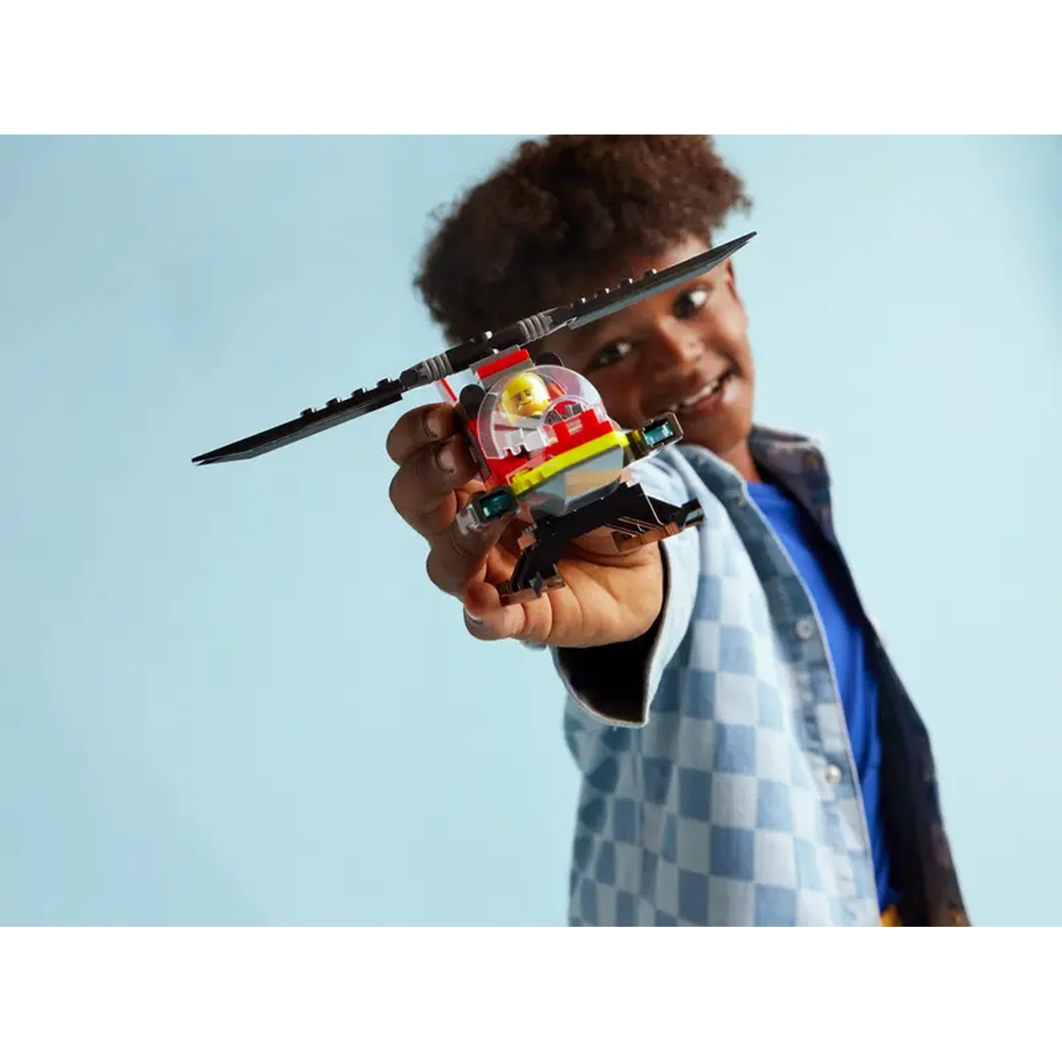 A smiling boy holds a Lego helicopter. The toy is black, red, yellow, and clear. He wears a blue shirt and jacket against a blue backdrop.