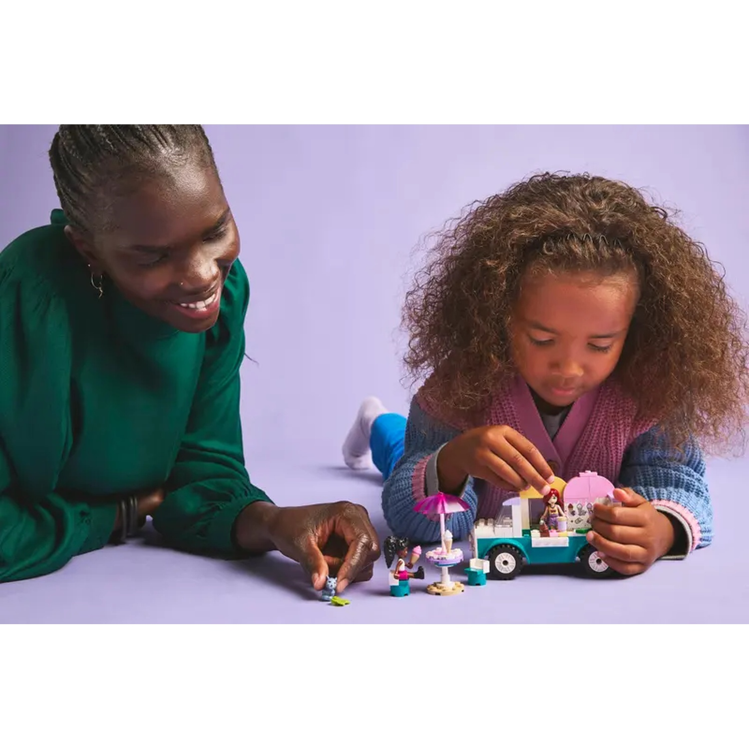 A woman and a girl play with a Lego ice cream truck. The truck is blue and white. They are on a purple surface.