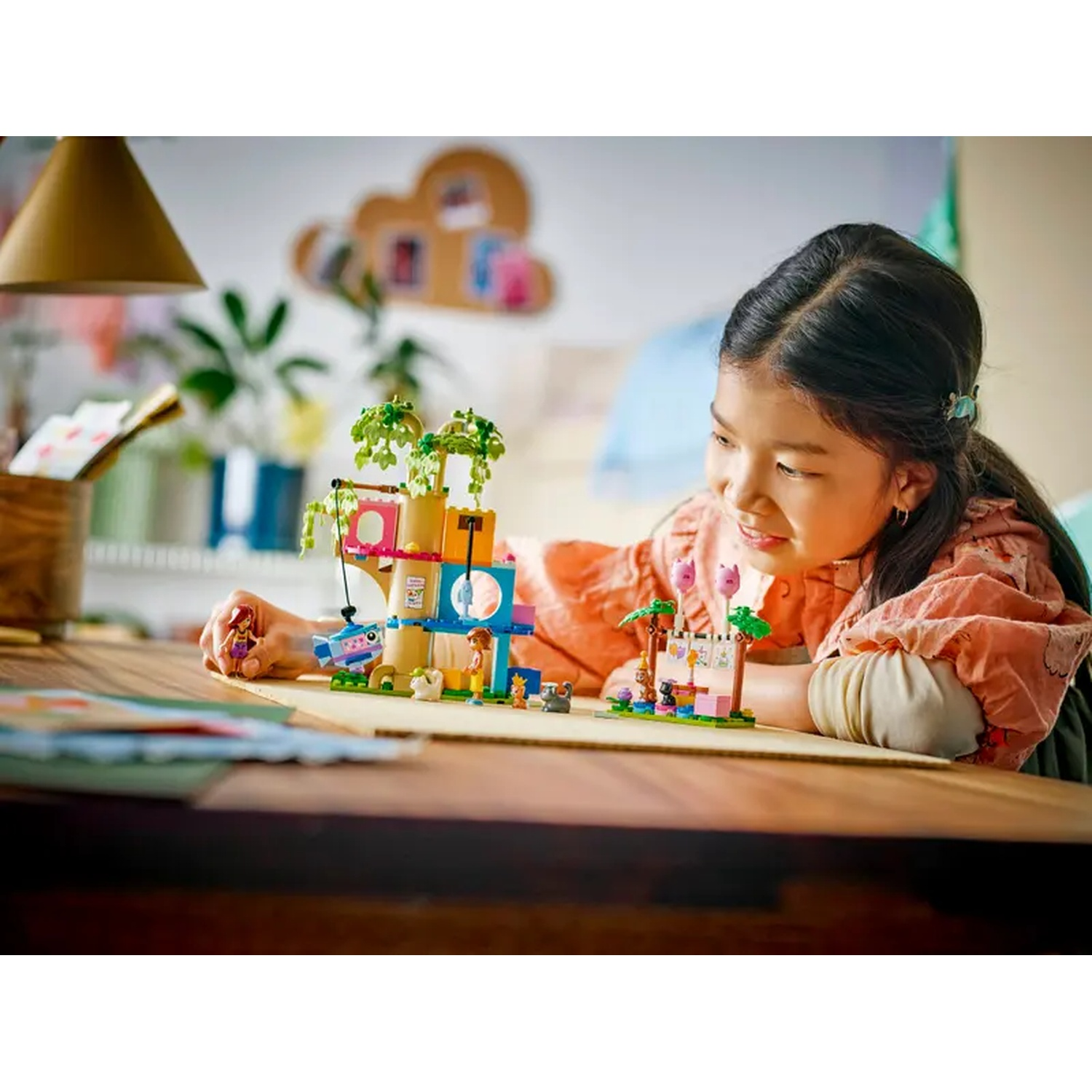A girl smiles while building a Lego set. The set shows a treehouse and a small display, both on a wooden table.