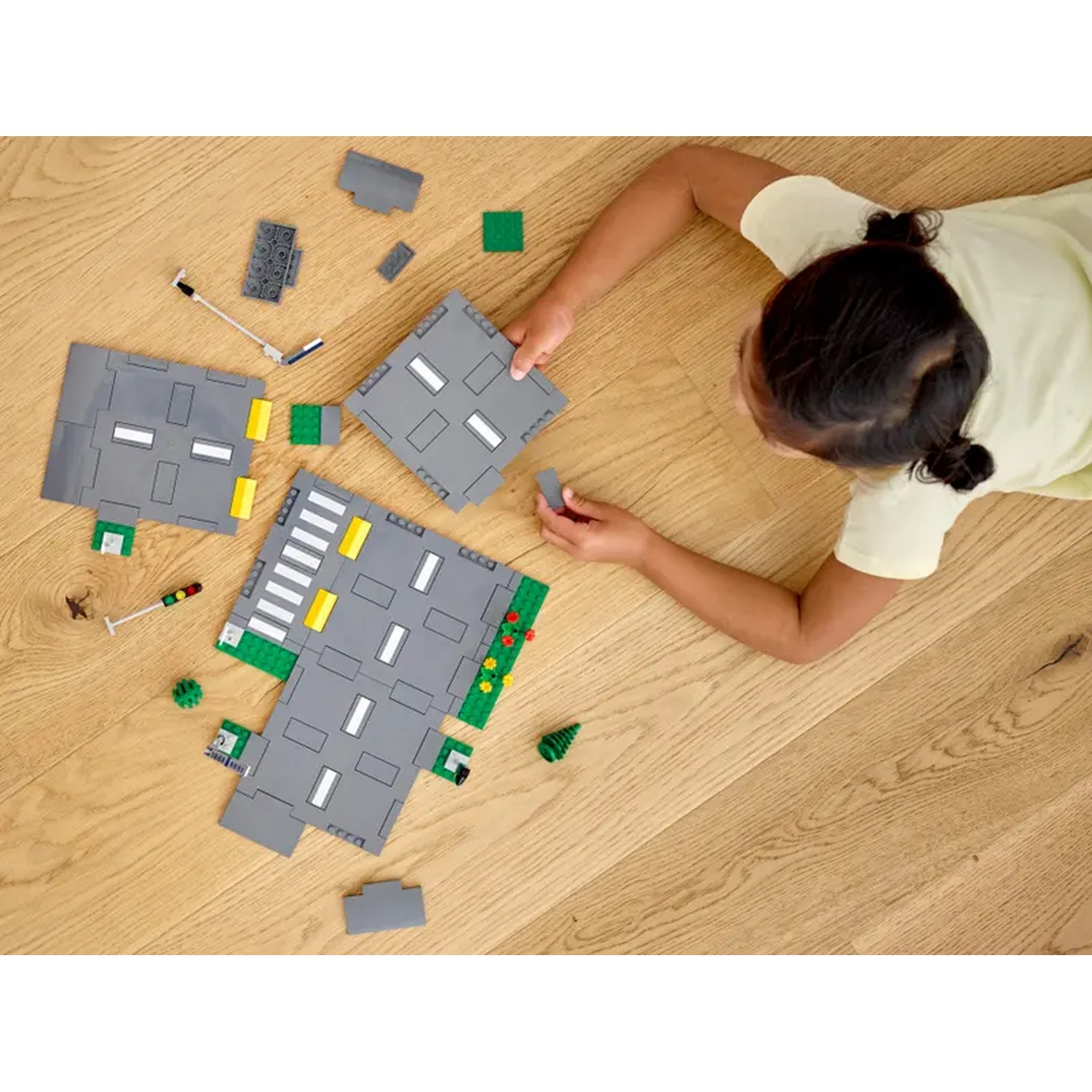 A child is building a road with toy blocks. The blocks are gray, green, and yellow on a wooden floor.