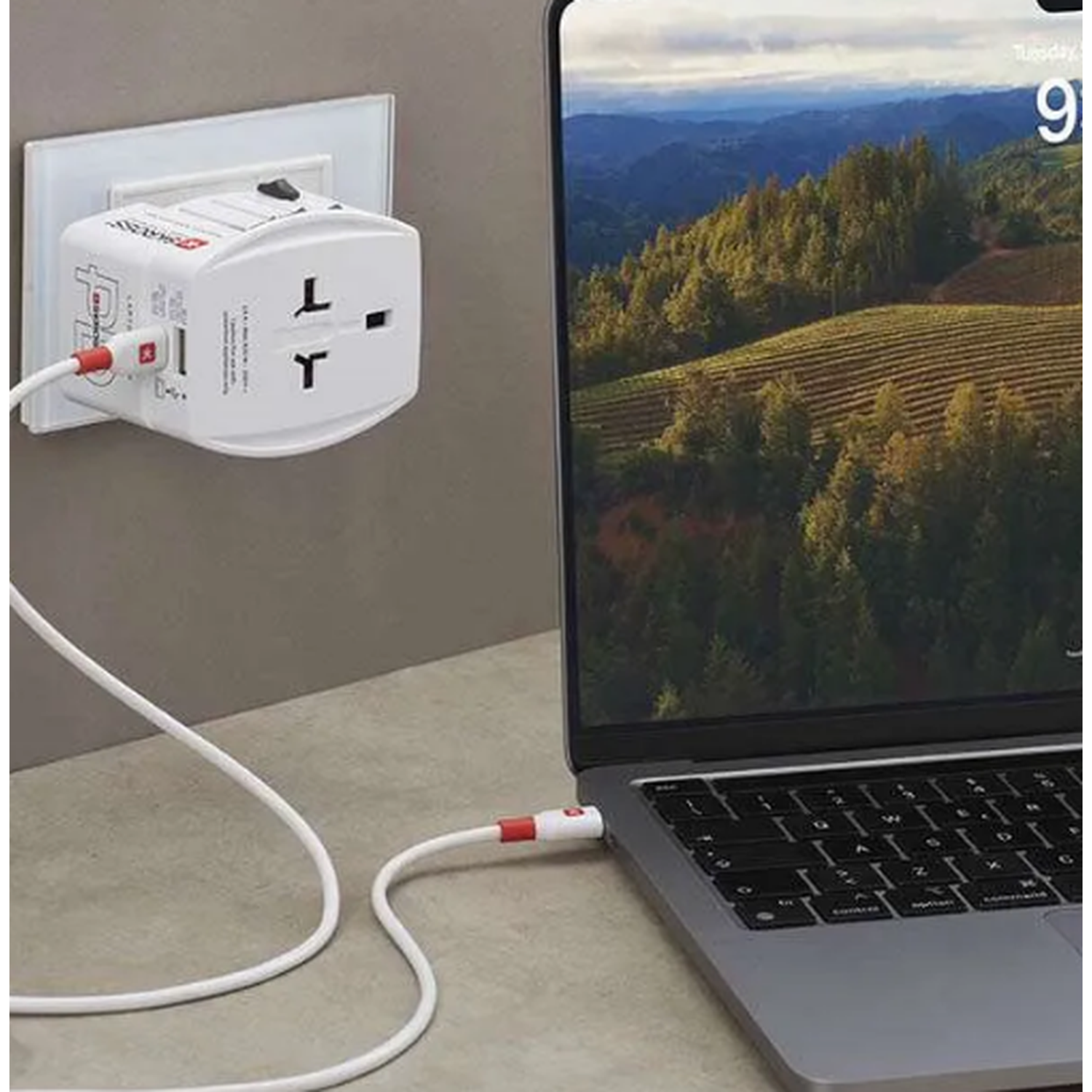 A white travel adapter plugged into a wall outlet, connected to a laptop with a charging cable.