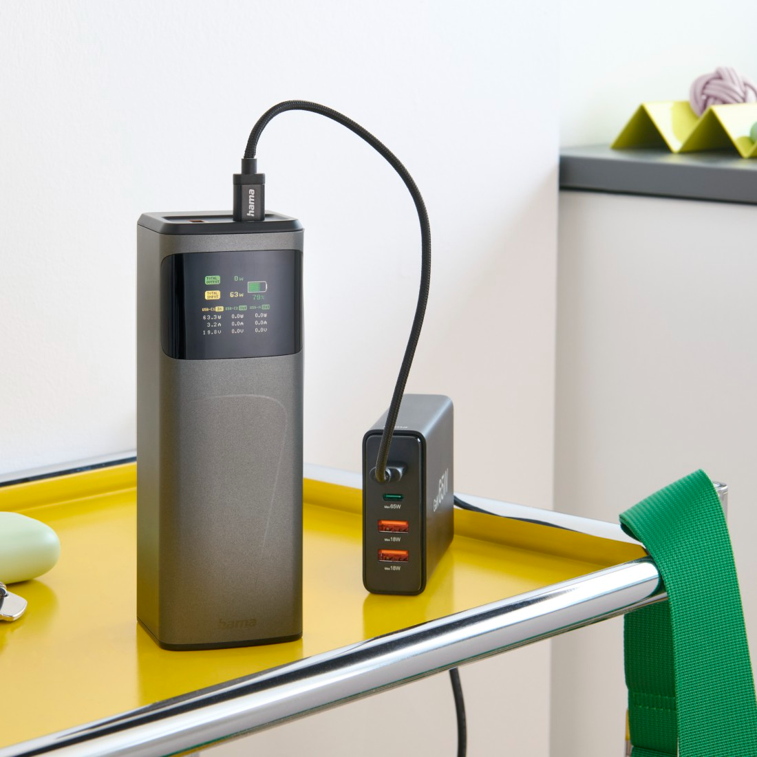 Egy szürke Hama power bank egy fekete töltőhöz van csatlakoztatva. Mindkettő egy sárga kocsin van. Egy zöld pánt lóg az oldalon.