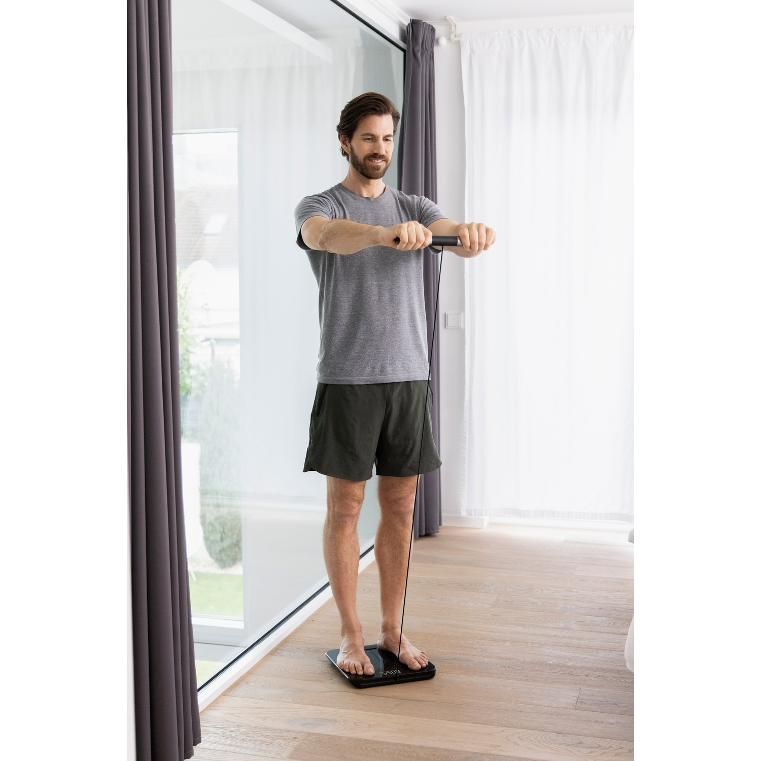 Een man staat op een weegschaal en houdt een apparaat vast. Hij draagt een grijs shirt en een korte broek. Grote ramen en gordijnen op de achtergrond.