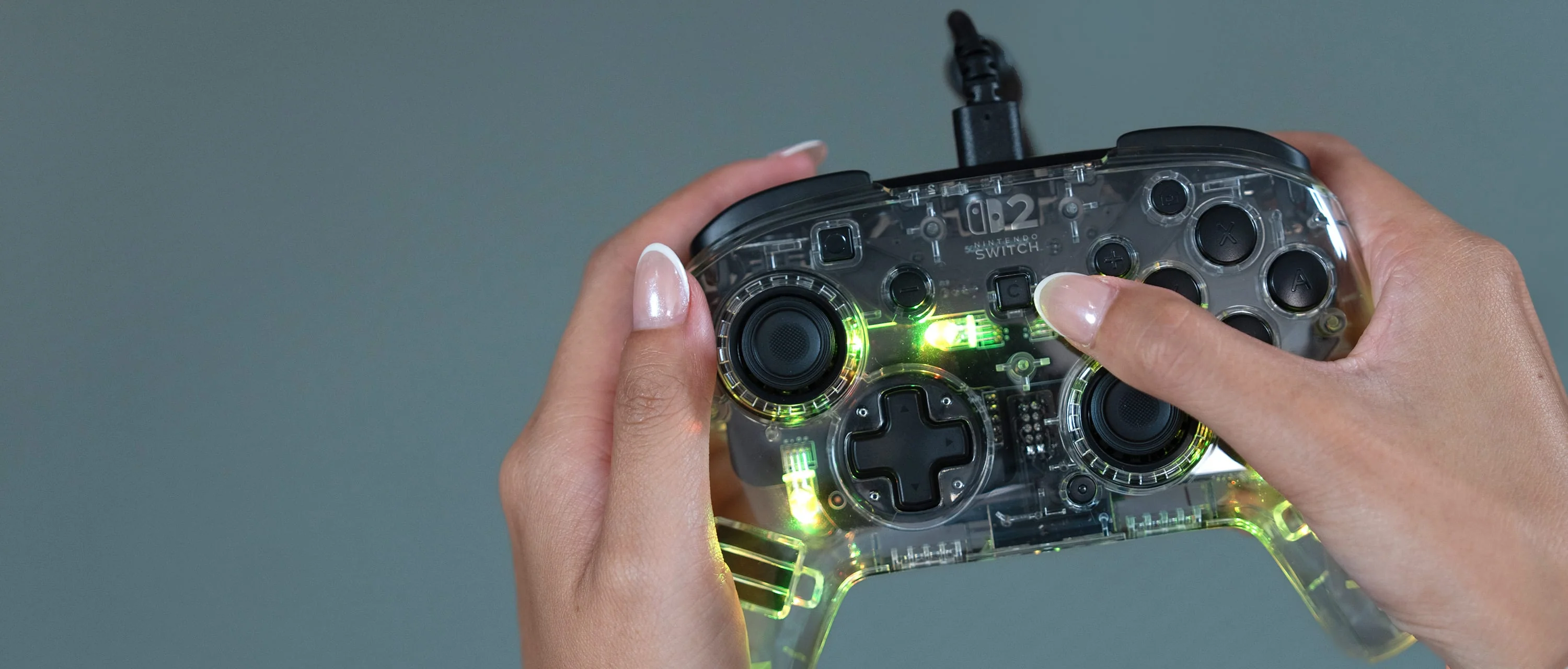 Hände halten einen durchsichtigen Nintendo Switch-Controller. Er hat ein schwarzes Kabel und die Knöpfe sind sichtbar. Der Hintergrund ist einfarbig grau.