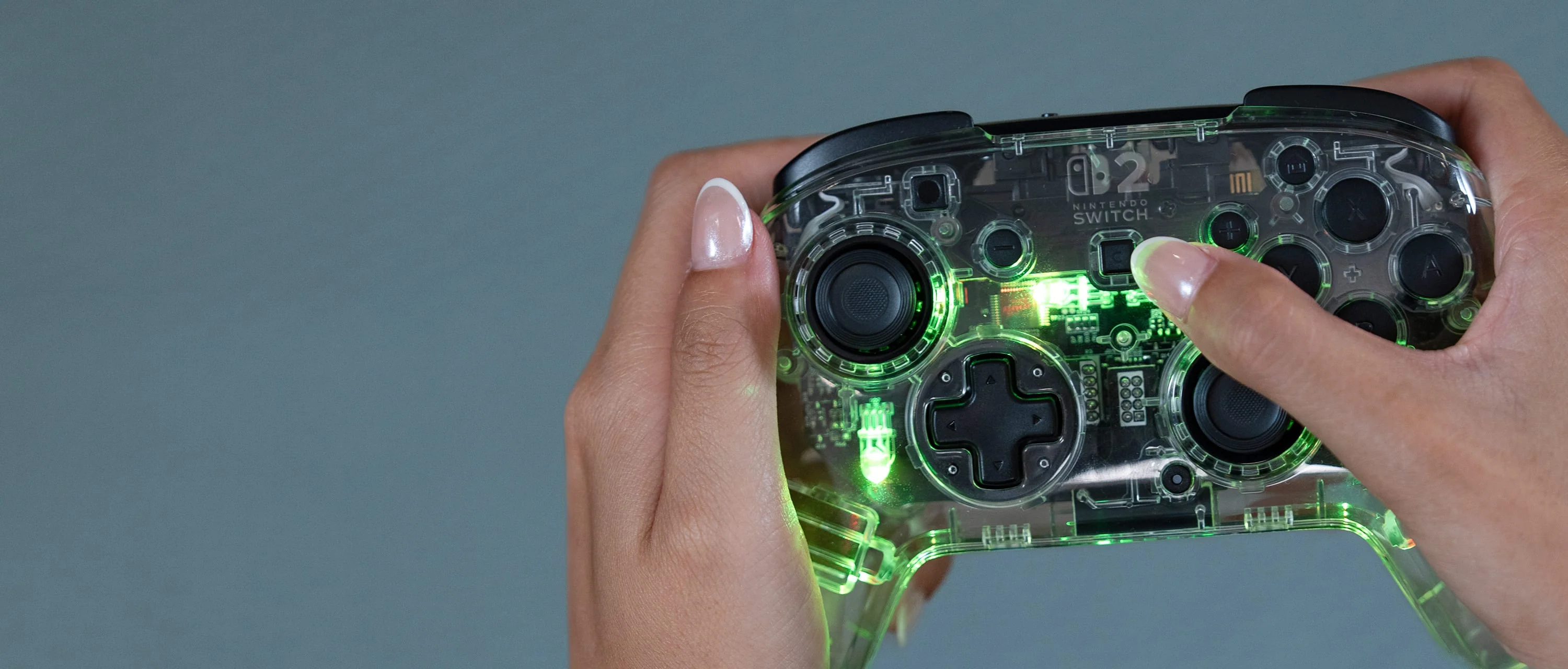 Hände halten einen transparenten Nintendo Switch-Controller. Grüne Lichter beleuchten das Innere. Der Hintergrund ist grau.