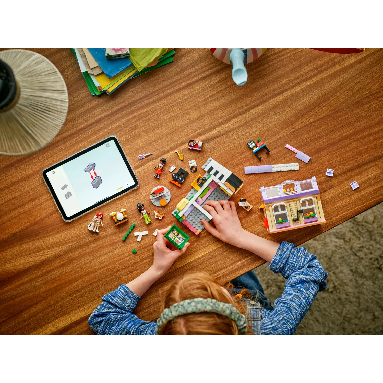 A child is building a Lego set on a wooden table. The set includes a house, figures, and musical instruments.
