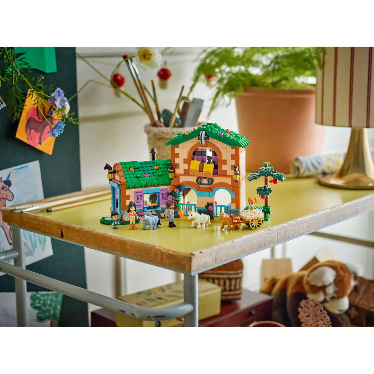 A Lego house with figures, horses, and a cart on a yellow table. Background includes drawings, plants, and a lamp.
