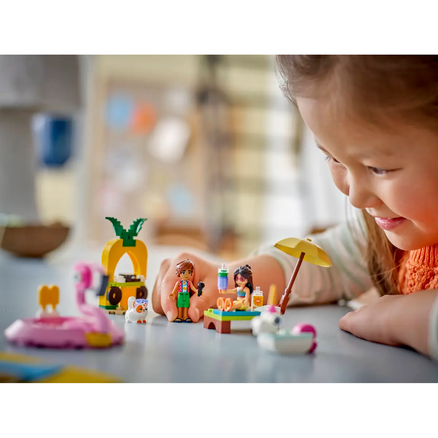 A child looks at a Lego set. The set includes a pineapple stand, flamingo, and figures on a table with a parasol.