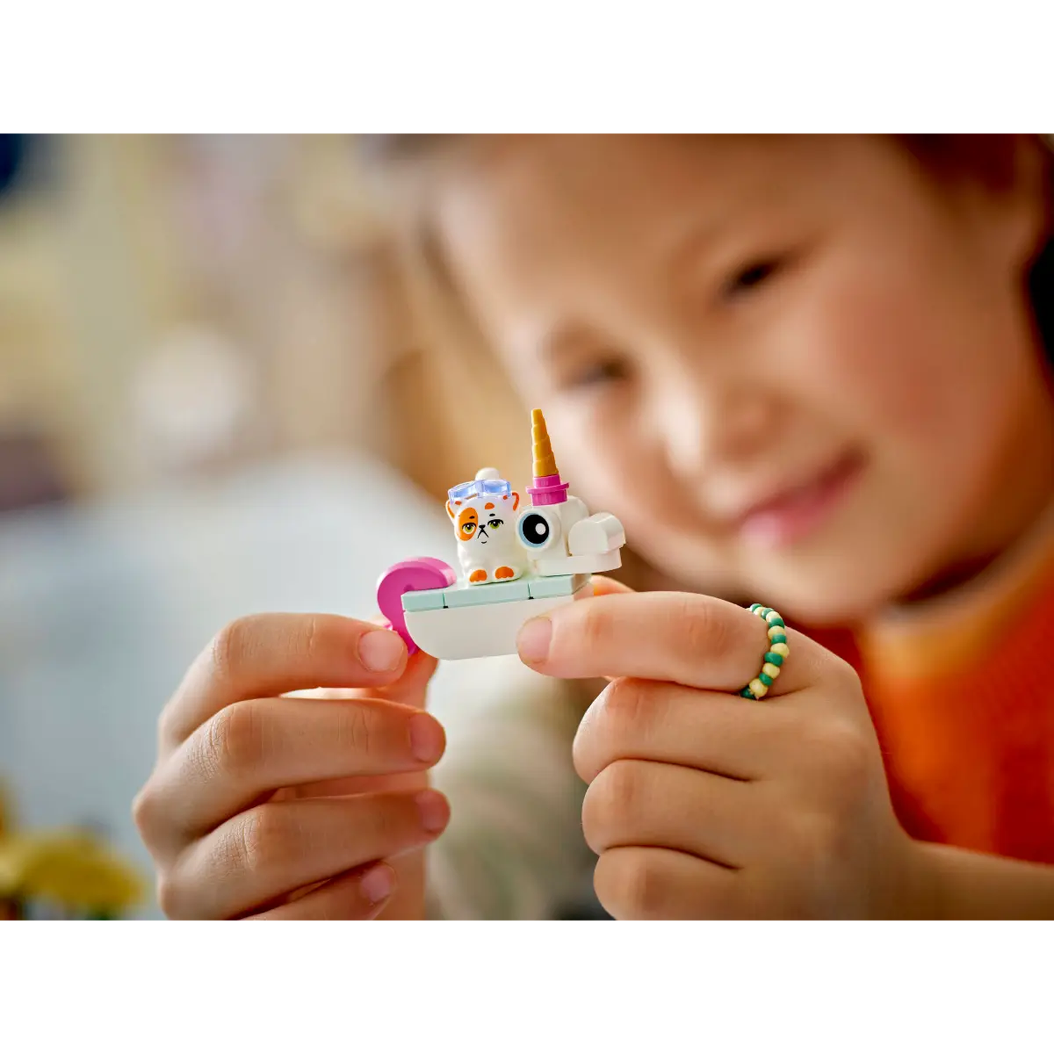 A child holds a toy cat-unicorn. The toy is white, pink, and orange. The child is wearing a beaded ring. Focus is on the toy.