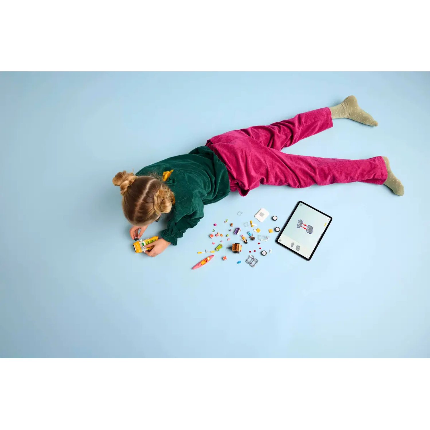 A child in green shirt and pink pants lies on a blue surface. Toys and a tablet are nearby. The child is assembling a toy.