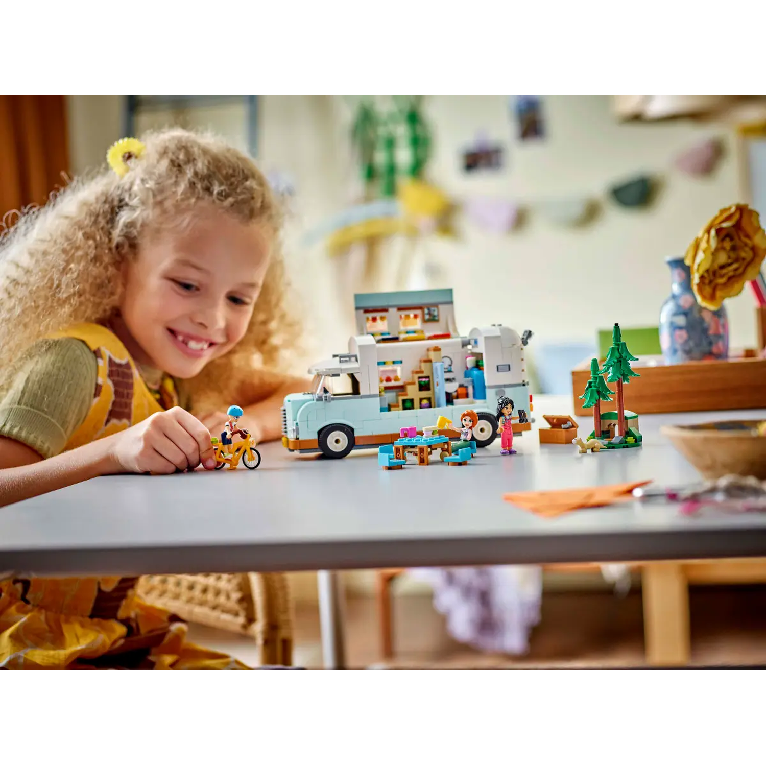 Een meisje glimlacht naar een LEGO-set met een foodtruck, figuren en accessoires op een tafel. Achtergrond bevat decor.