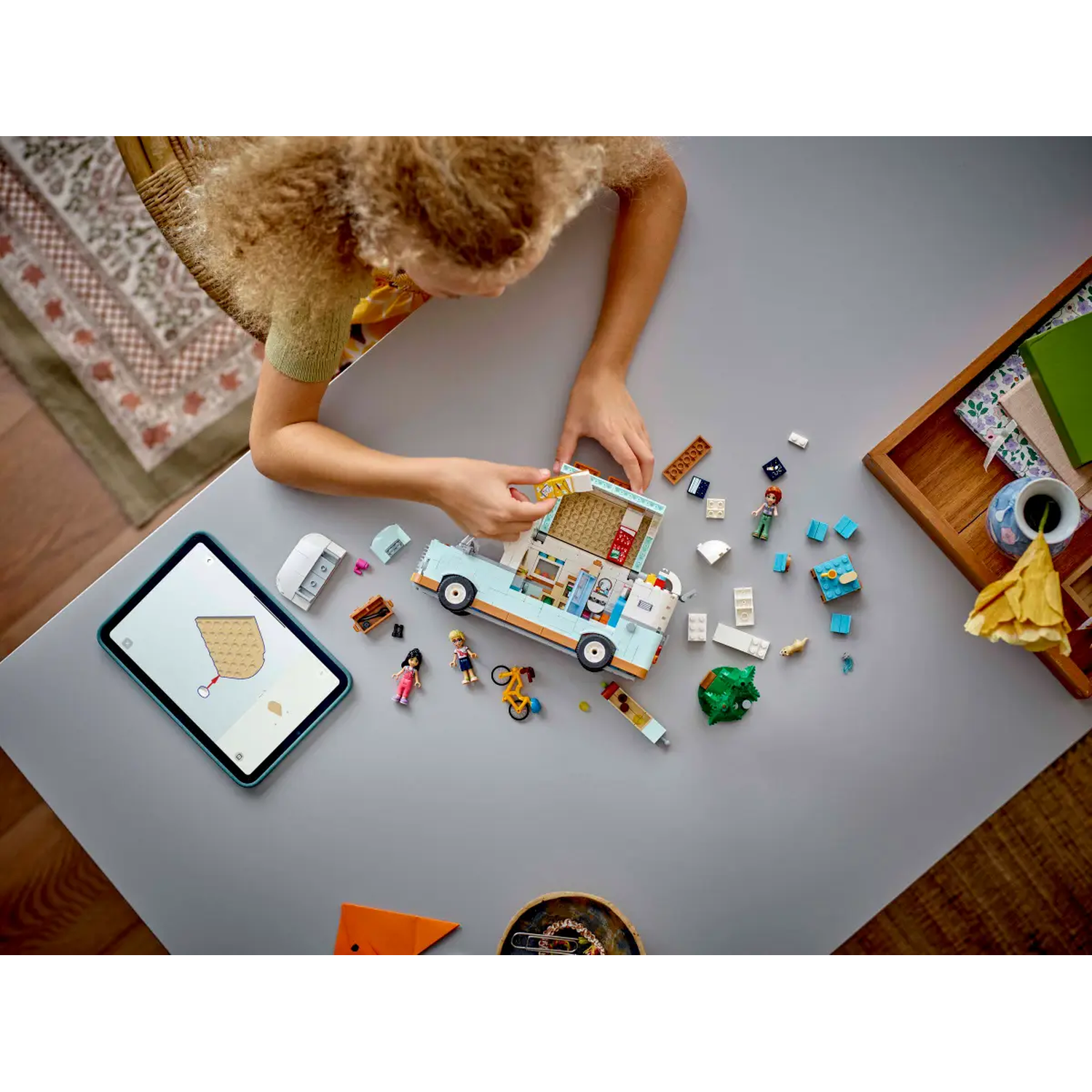 Een kind assembleert een speelgoedcamper op een grijze tafel. Legostenen en minifiguren liggen verspreid. Een tablet toont instructies.