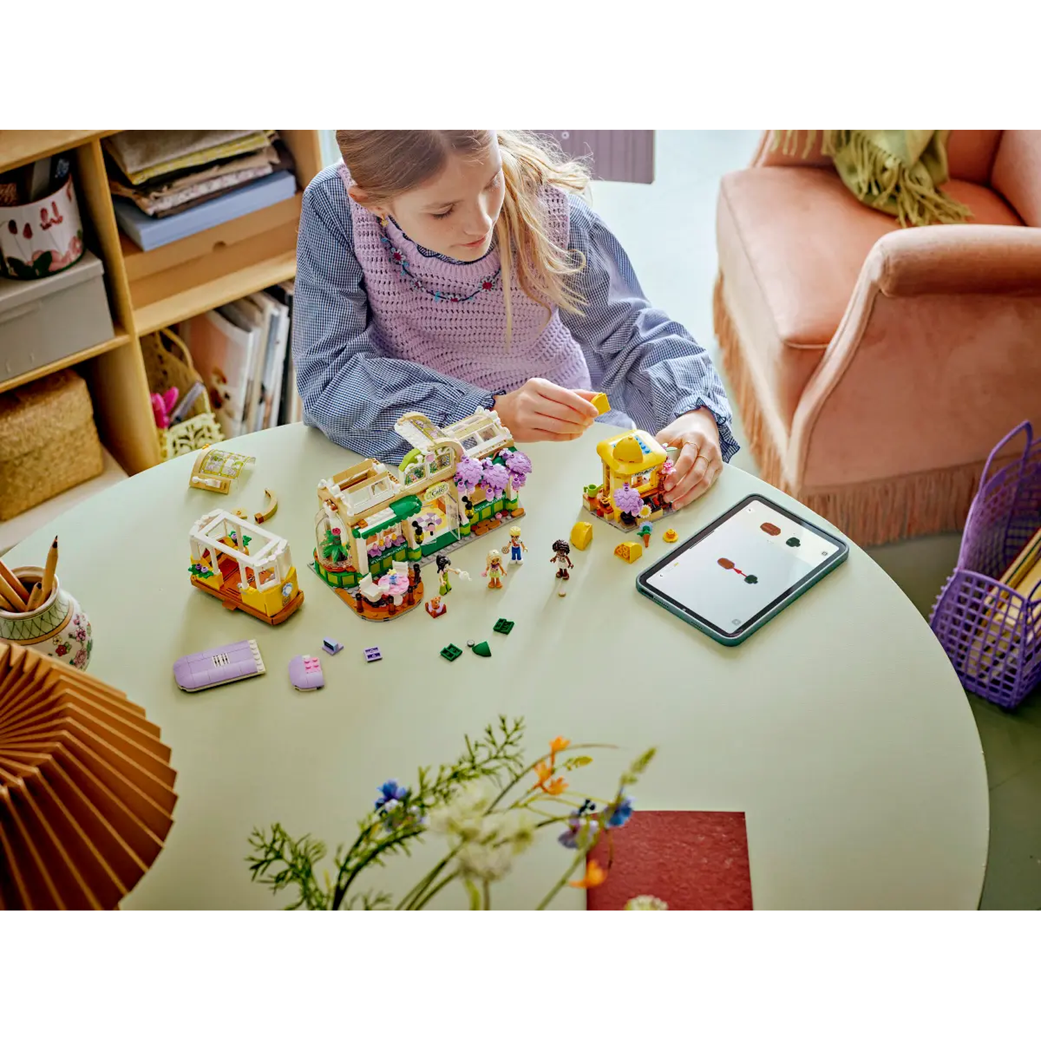 A girl plays with Lego sets on a table. Lego figures and buildings are arranged. A tablet is on the table.