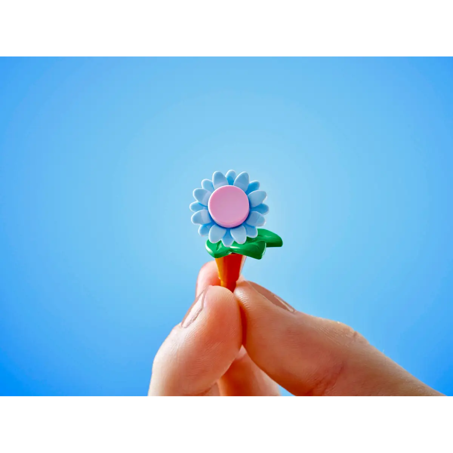A hand holding a toy flower against a blue background. The flower is blue and pink, with green leaves.