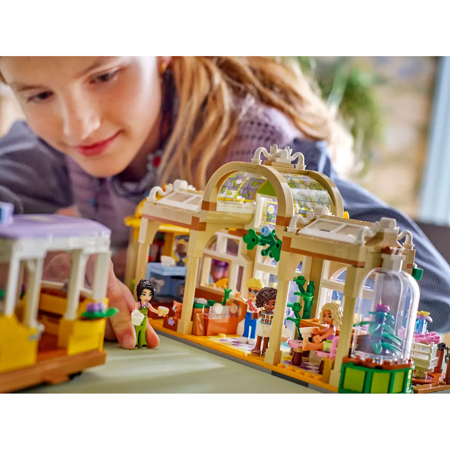 A girl looks at a Lego set with a greenhouse and figures. The set includes a yellow tram and miniature figures.
