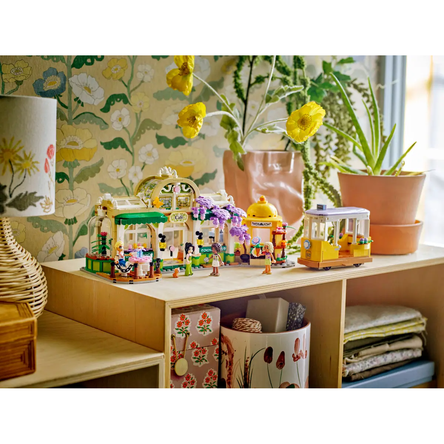 A Lego set of a cafe and tram on a wooden shelf, with flowers and plants in the background. Colorful and detailed.