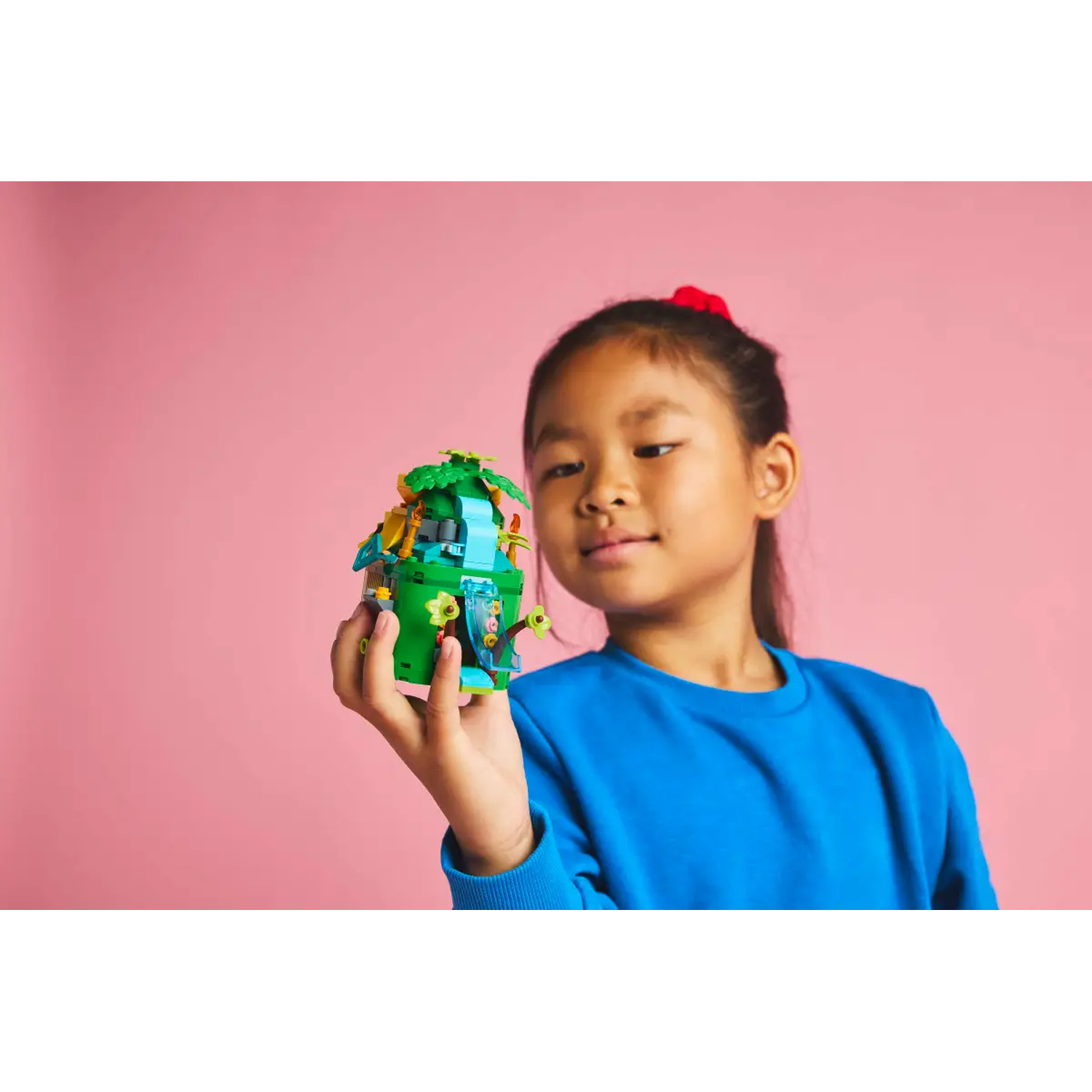 A girl holds a green Lego building with a pink background. She wears a blue shirt and has a red hair tie.