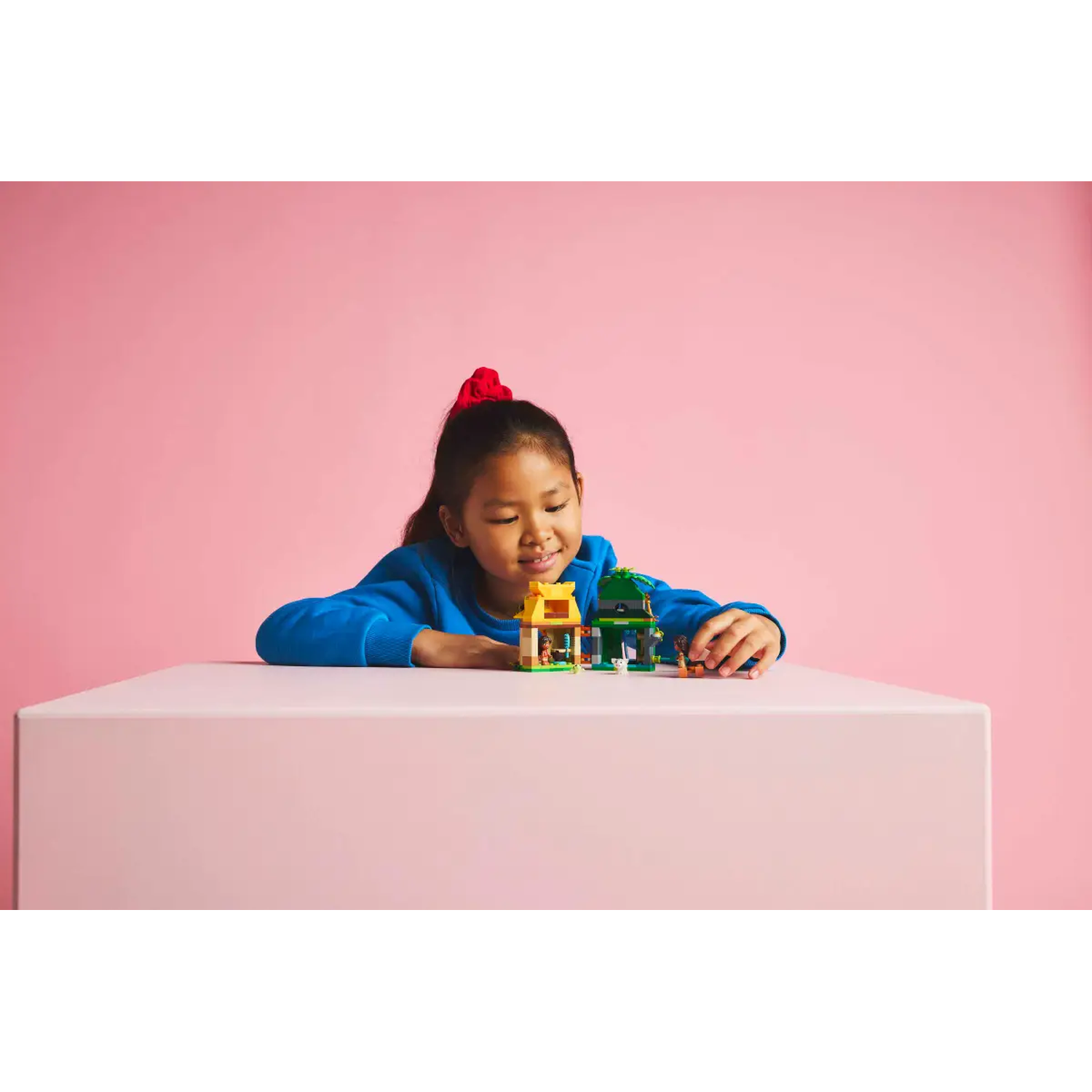 A young girl with a red bow plays with Lego houses on a pink table. The background is pink.