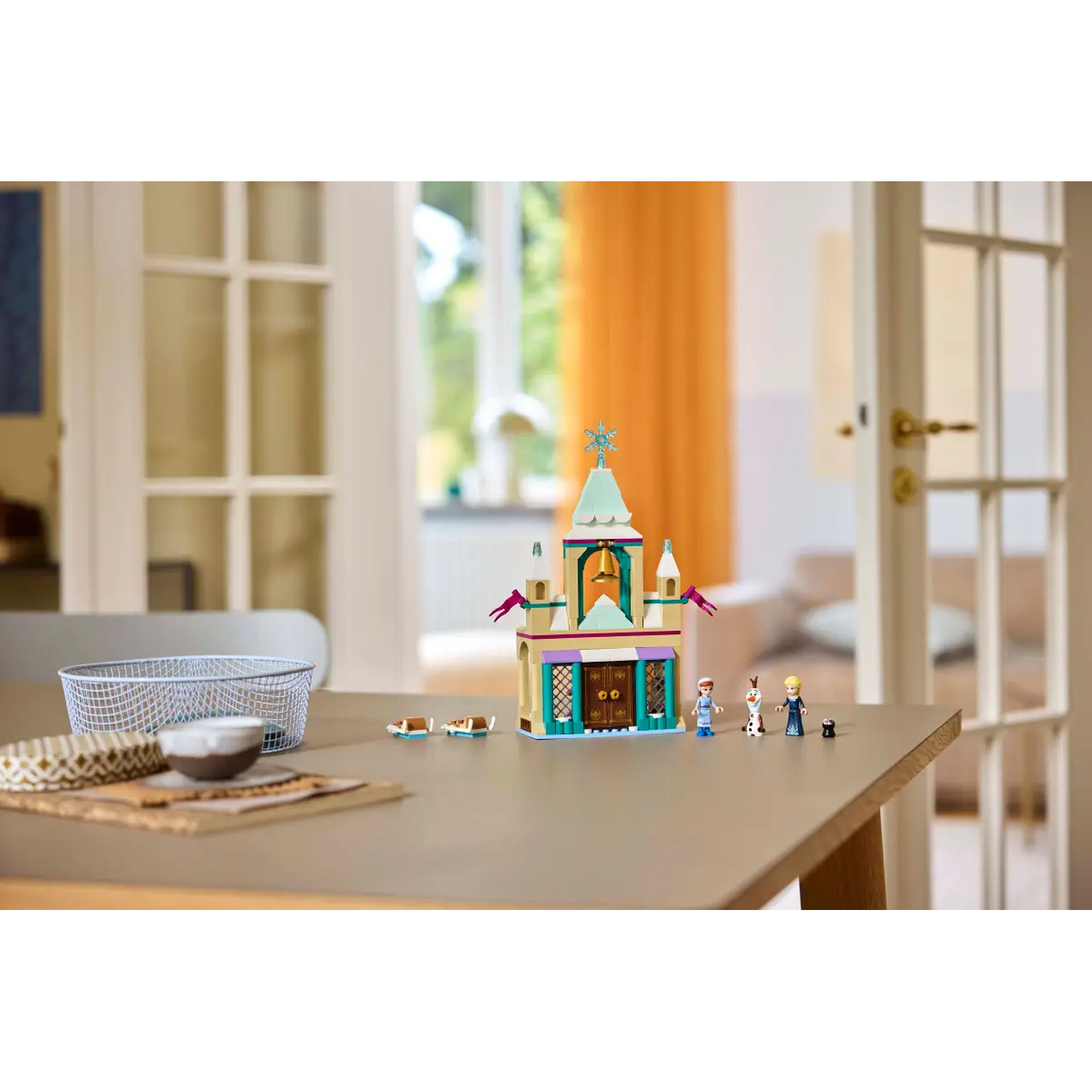 A Lego castle is on a table. Three Lego figures and Olaf are near it. A basket, bowl and patterned mat are on the table.