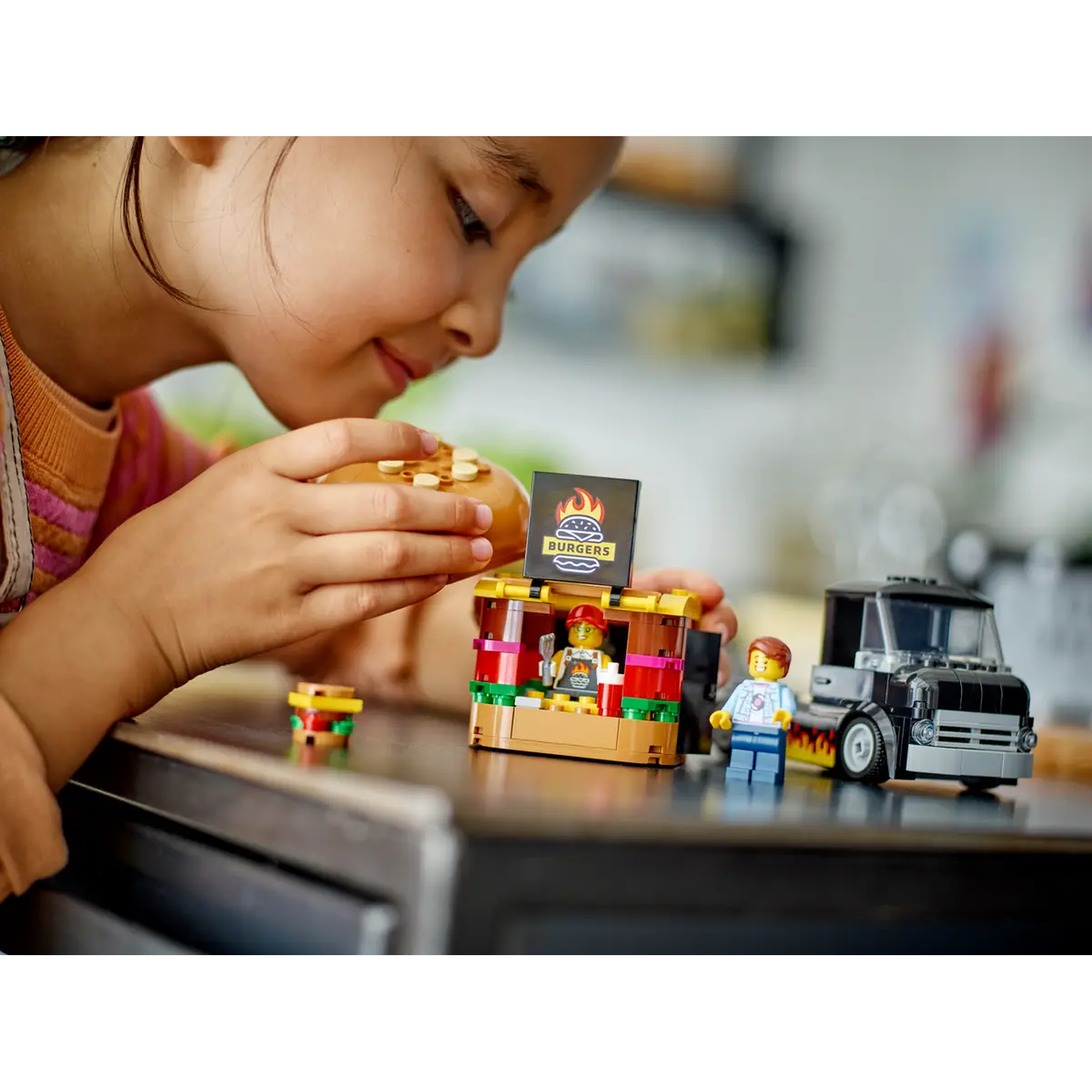 A child is playing with a Lego set, focused on a burger. A food truck and burger stand are also in the scene.