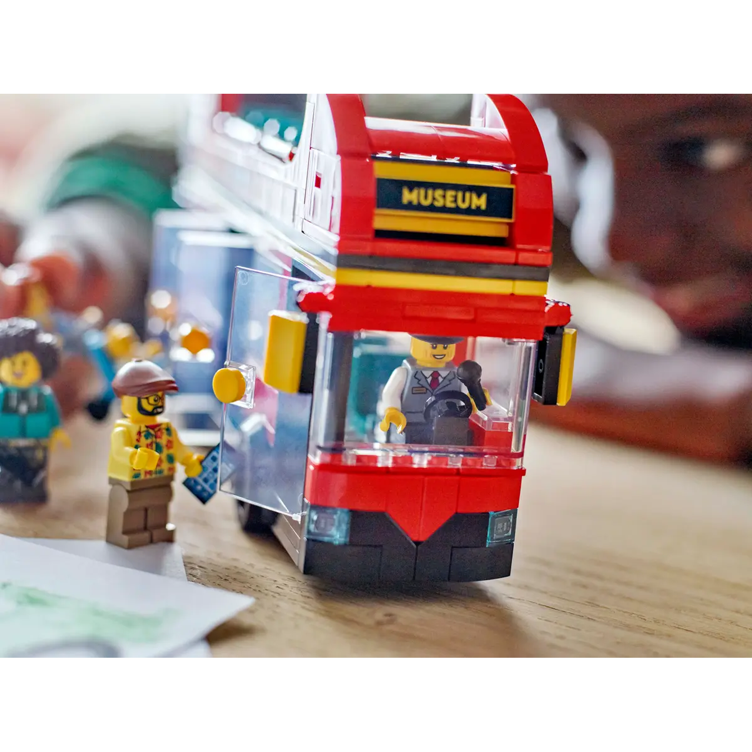 A Lego double-decker bus with a driver, and people. The bus is red with a museum sign. A person is holding a blue ticket.