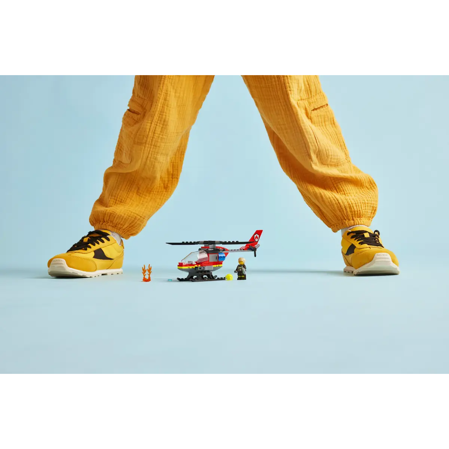 A person in yellow pants and shoes stands over a Lego helicopter and minifigures. Background is light blue.
