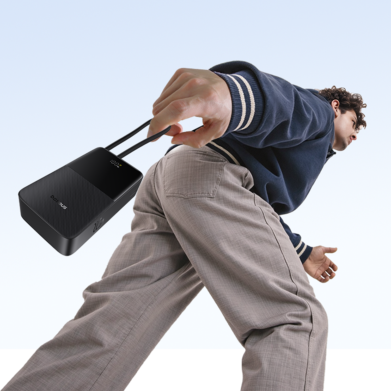 Un hombre sostiene un banco de energía negro, con una chaqueta azul y pantalones grises. Fondo del cielo. El banco muestra 100%.