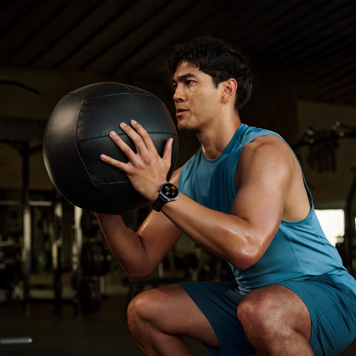 Un hombre en un gimnasio sostiene un balón medicinal negro. Viste camiseta sin mangas y pantalones cortos azules, y un reloj inteligente. Está en cuclillas.