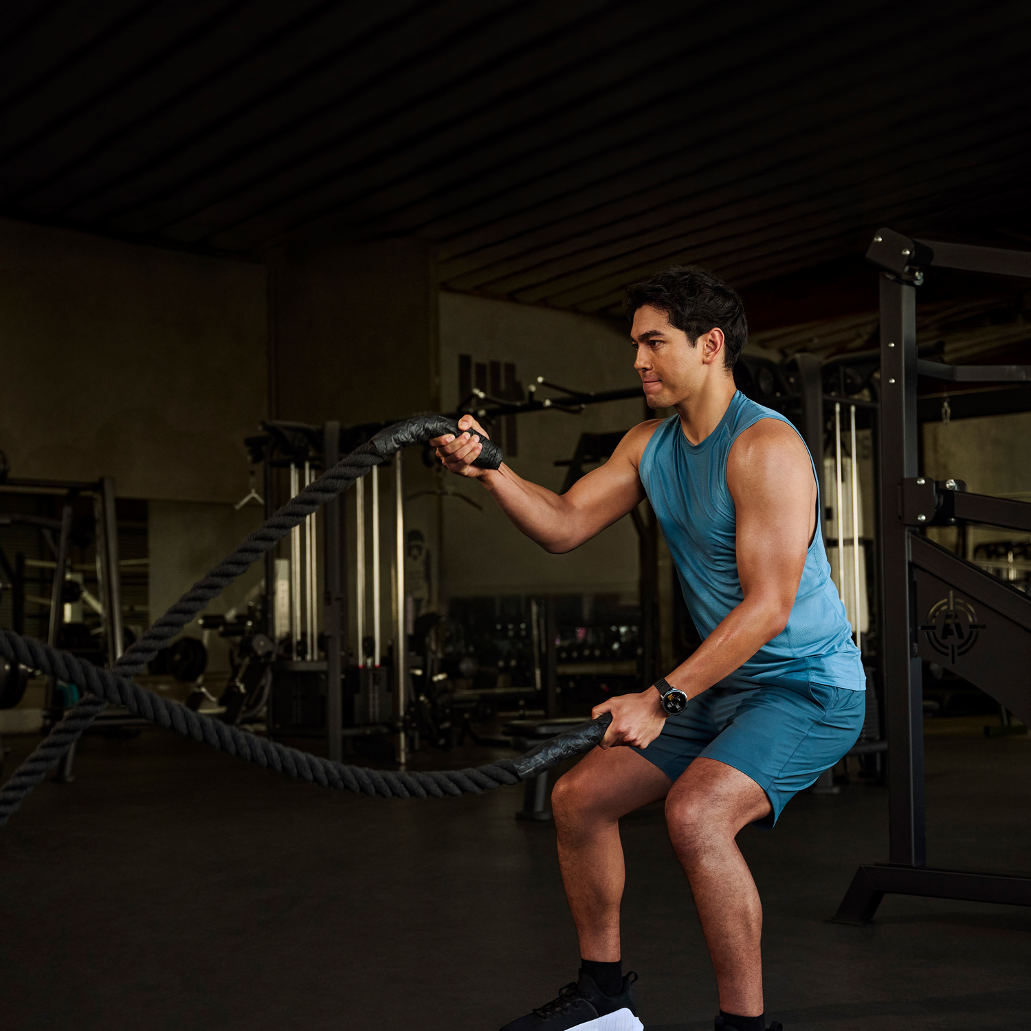 Un hombre en un gimnasio está haciendo ejercicio con cuerdas de batalla. Viste ropa deportiva azul. Hay equipos de gimnasio en el fondo.