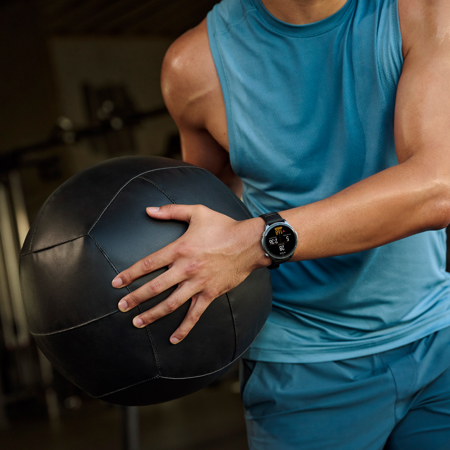 Una persona con una camiseta sin mangas azul sostiene un balón medicinal negro. Un reloj inteligente muestra estadísticas. La escena es un gimnasio.